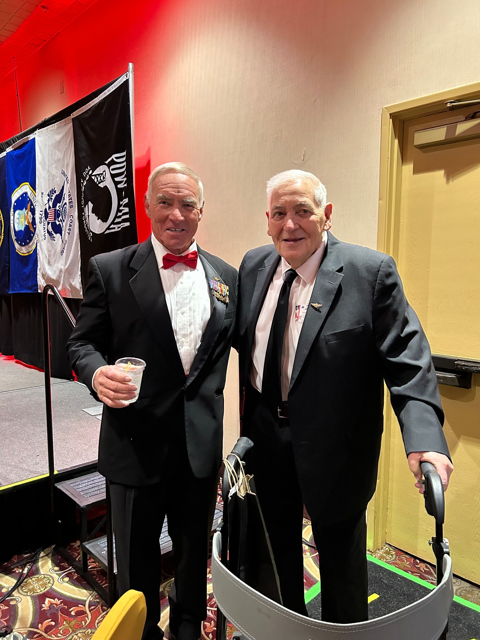 Two men in tuxedos pose at an event, one holding a drink, with flags and a doorway behind them.