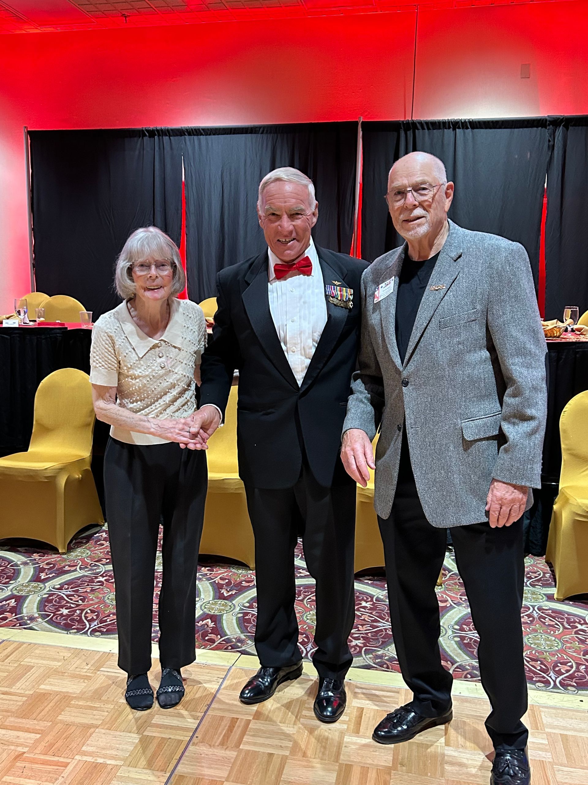 Three people posing indoors at a formal event, with a red backdrop and patterned carpet.