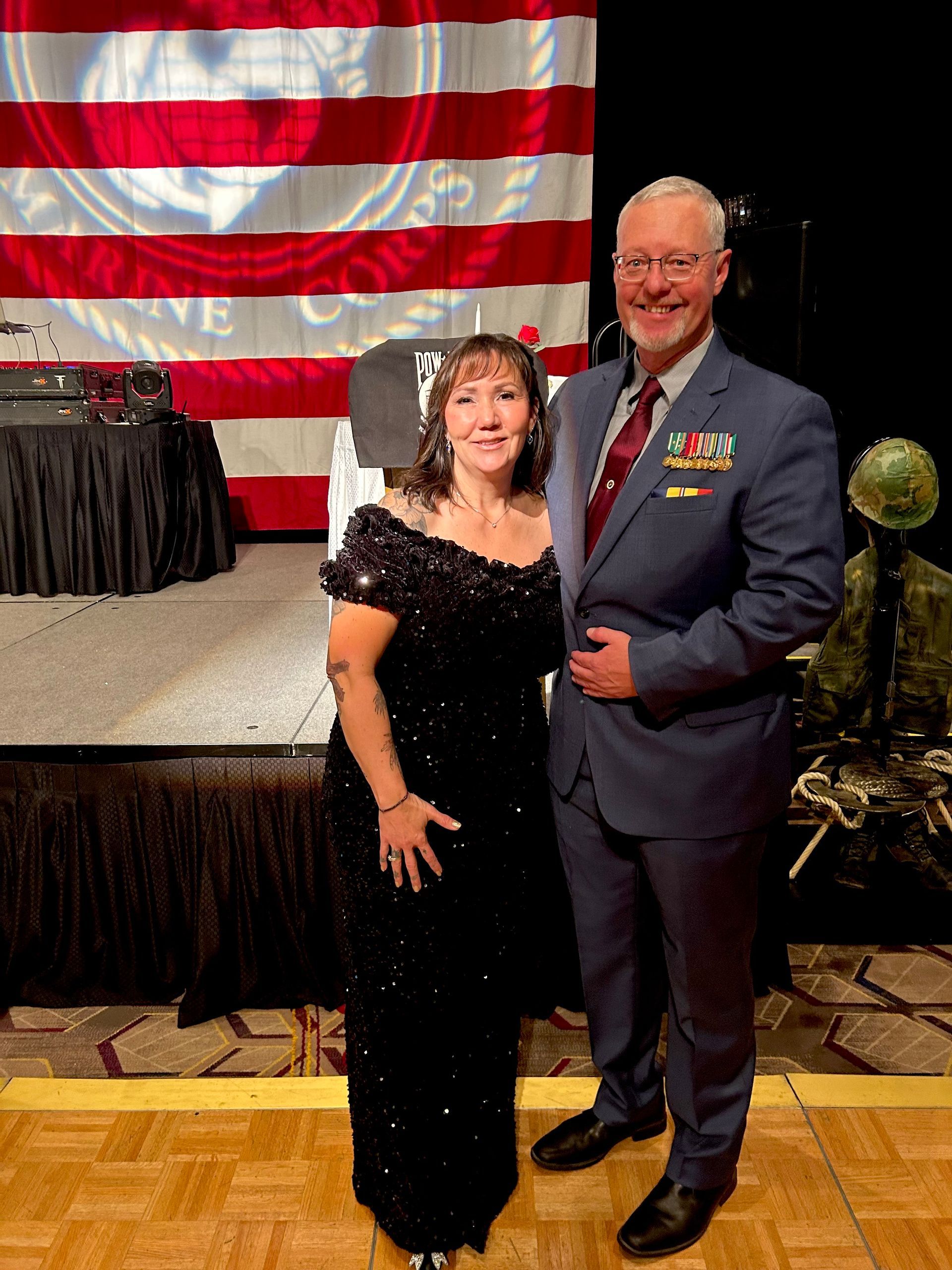 Woman in black gown and man in dress uniform posing on a stage with an American flag backdrop.