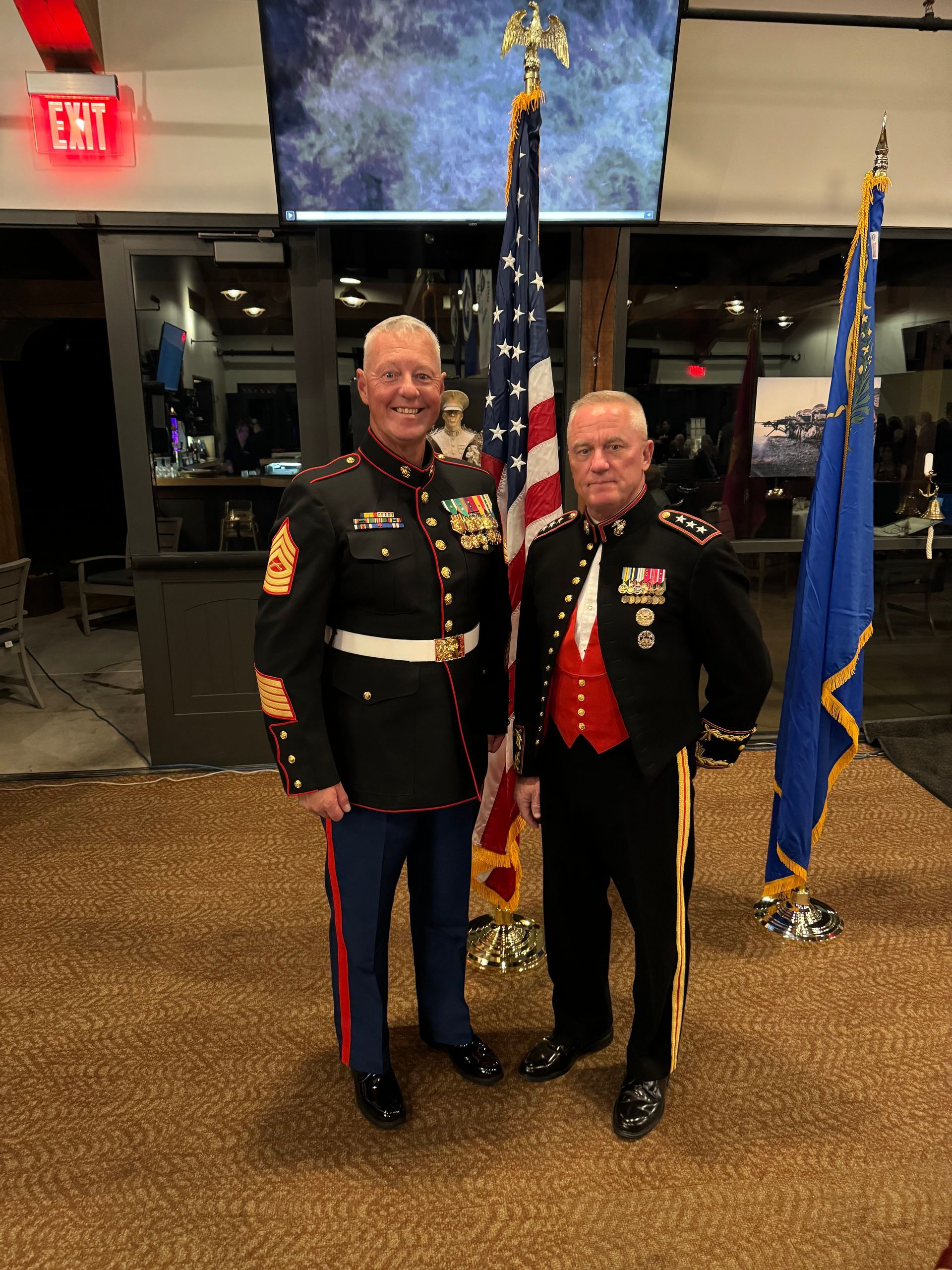 Two men in decorated military dress uniforms stand indoors beside U.S. and blue flags.