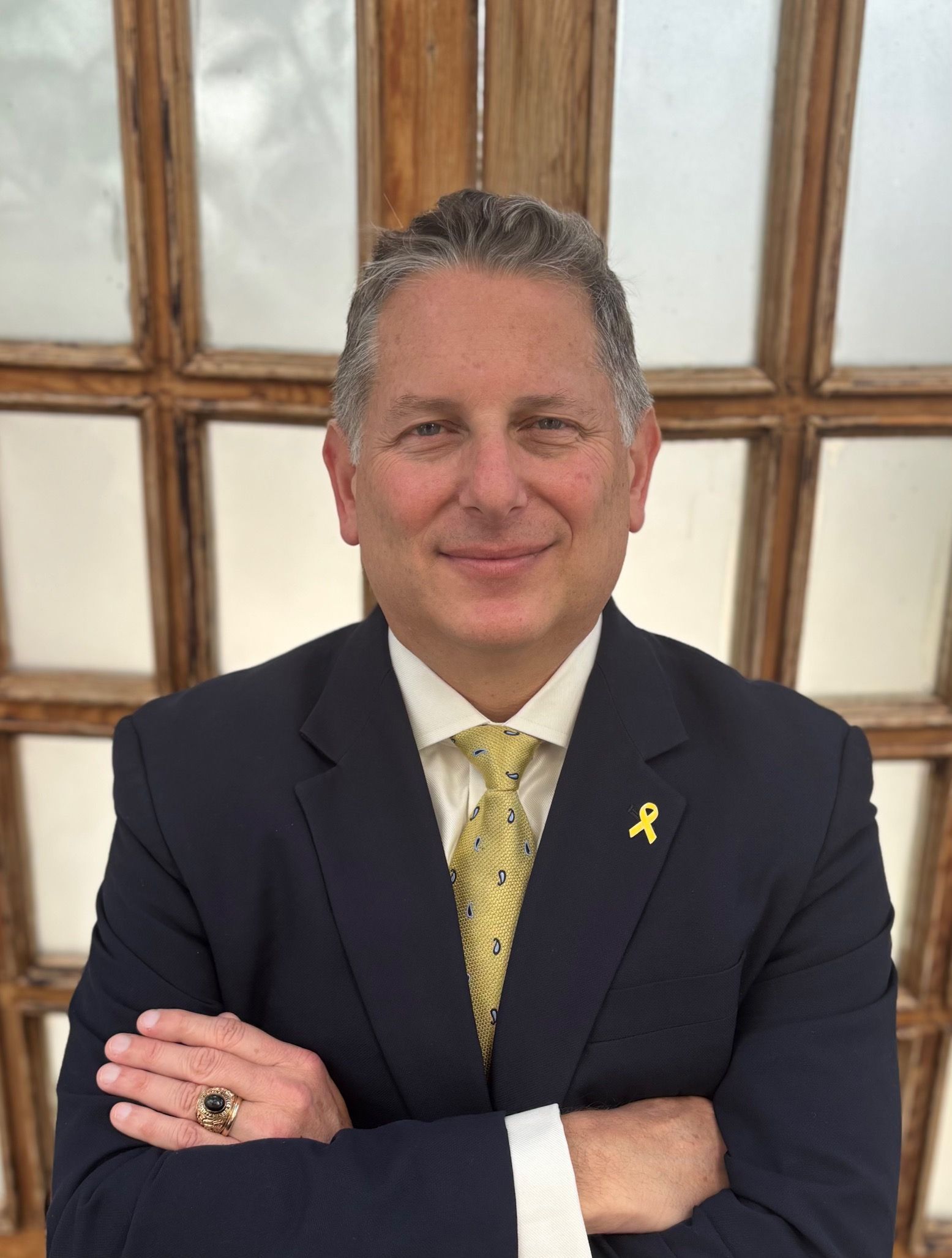 Man in dark suit with yellow tie and lapel ribbon, arms crossed, standing before frosted windows.