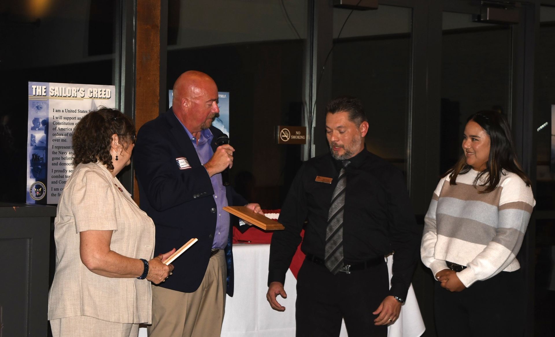 Four people stand indoors at a formal event while one man speaks into a microphone and holds a plaque.