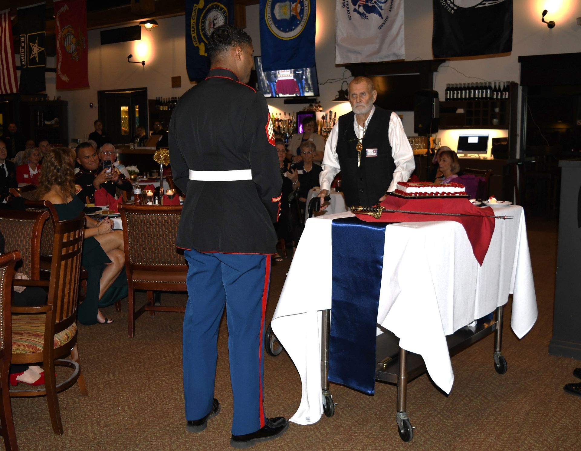 Server in black jacket and blue pants stands by a table with a red-and-white tablecloth in a restaurant.