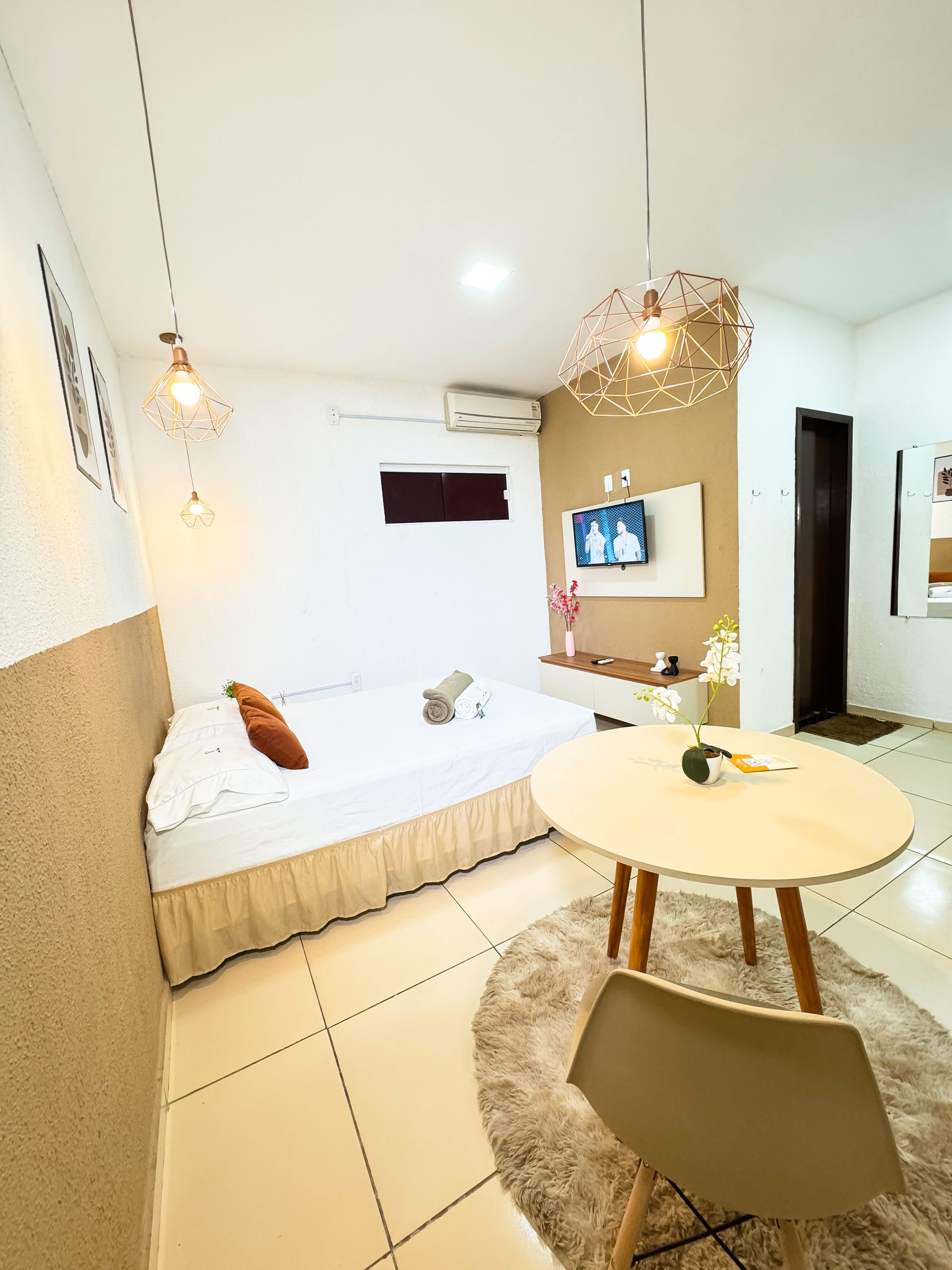 Apartamento estúdio aconchegante com cama, mesa, TV e iluminação decorativa.