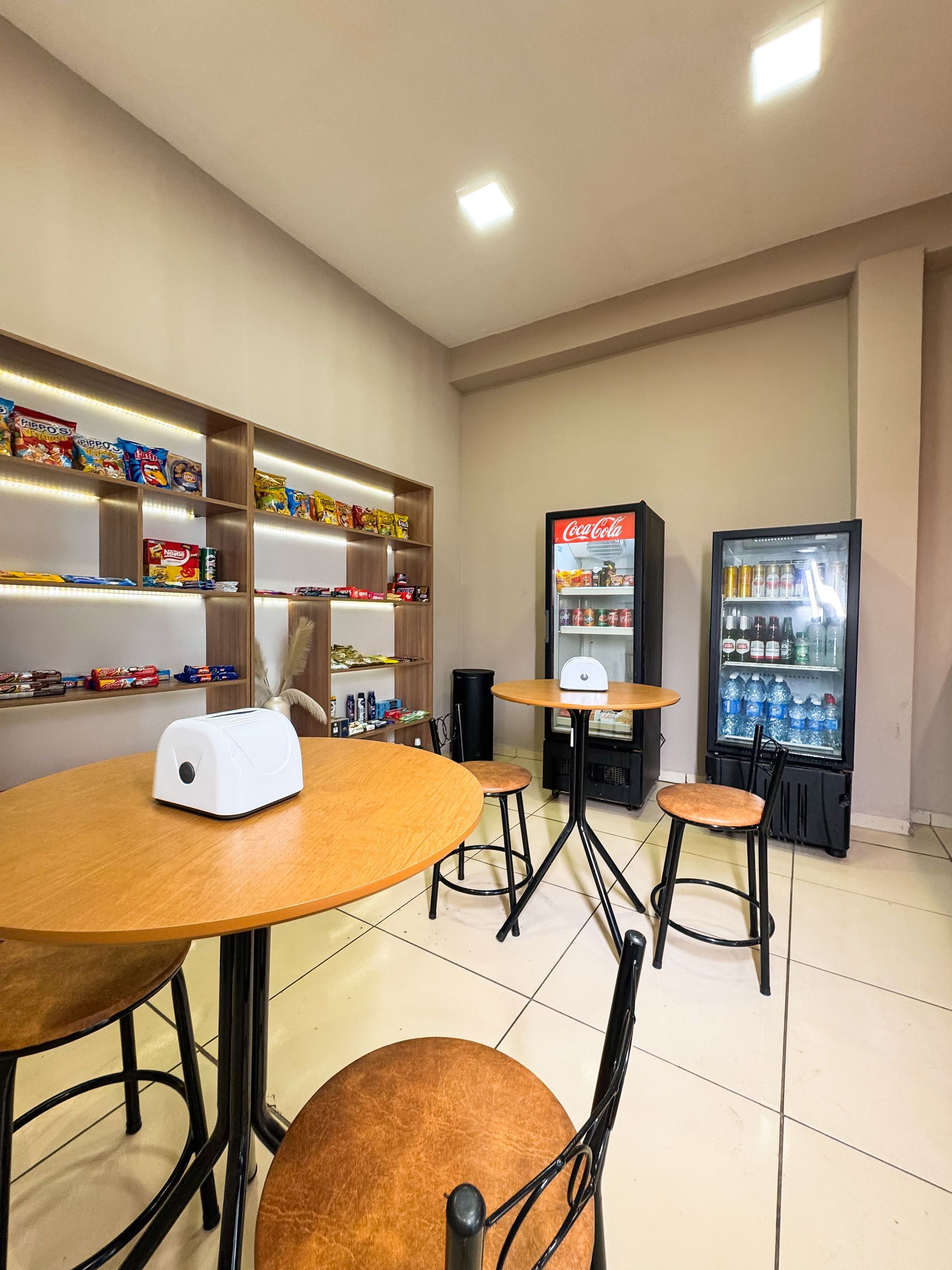Interior de uma pequena loja com prateleiras de lanches, geladeiras e mesas redondas com bancos.