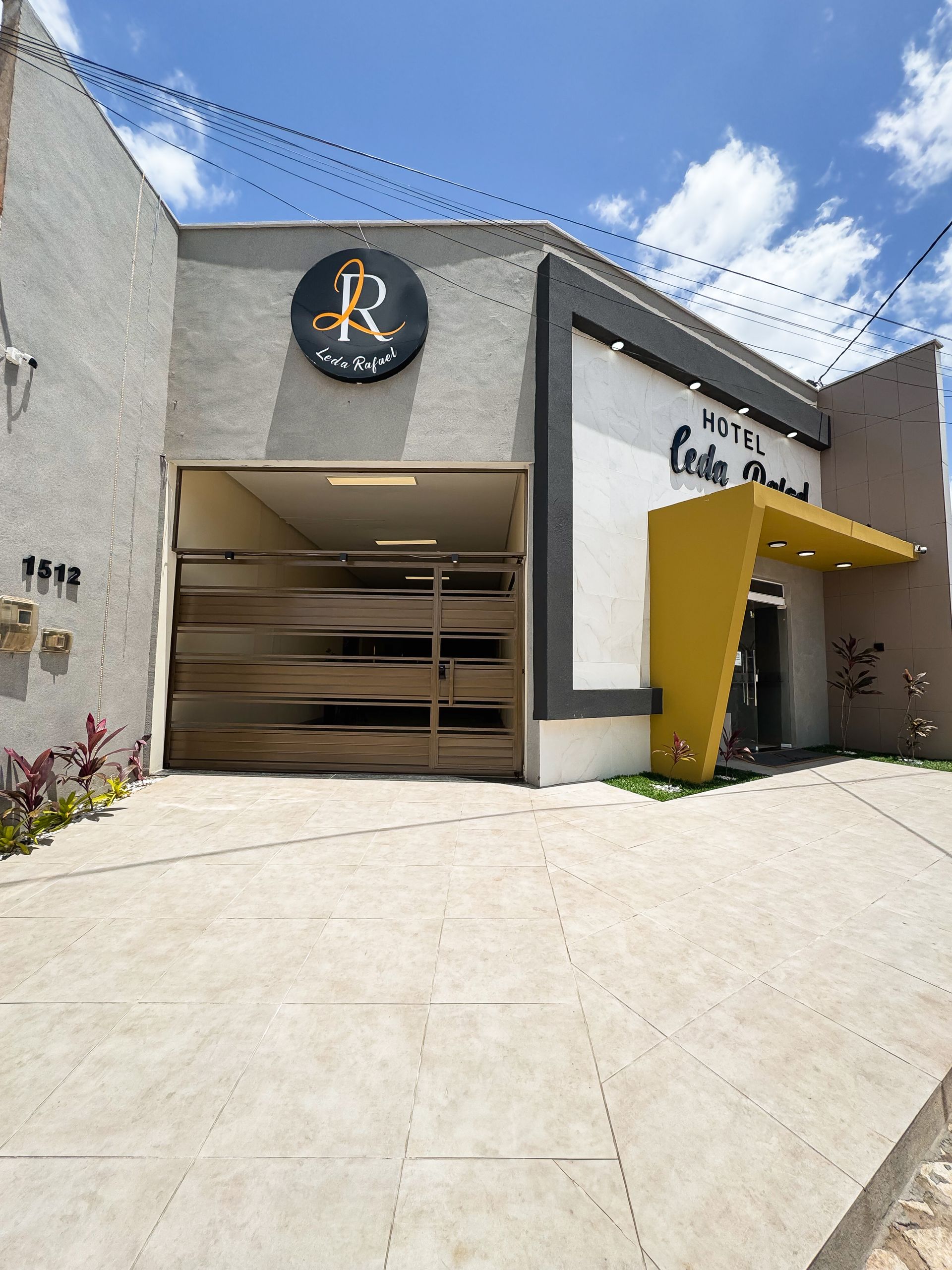 Exterior do edifício com fachada cinza, portão metálico em arco e entrada amarela. A placa diz 