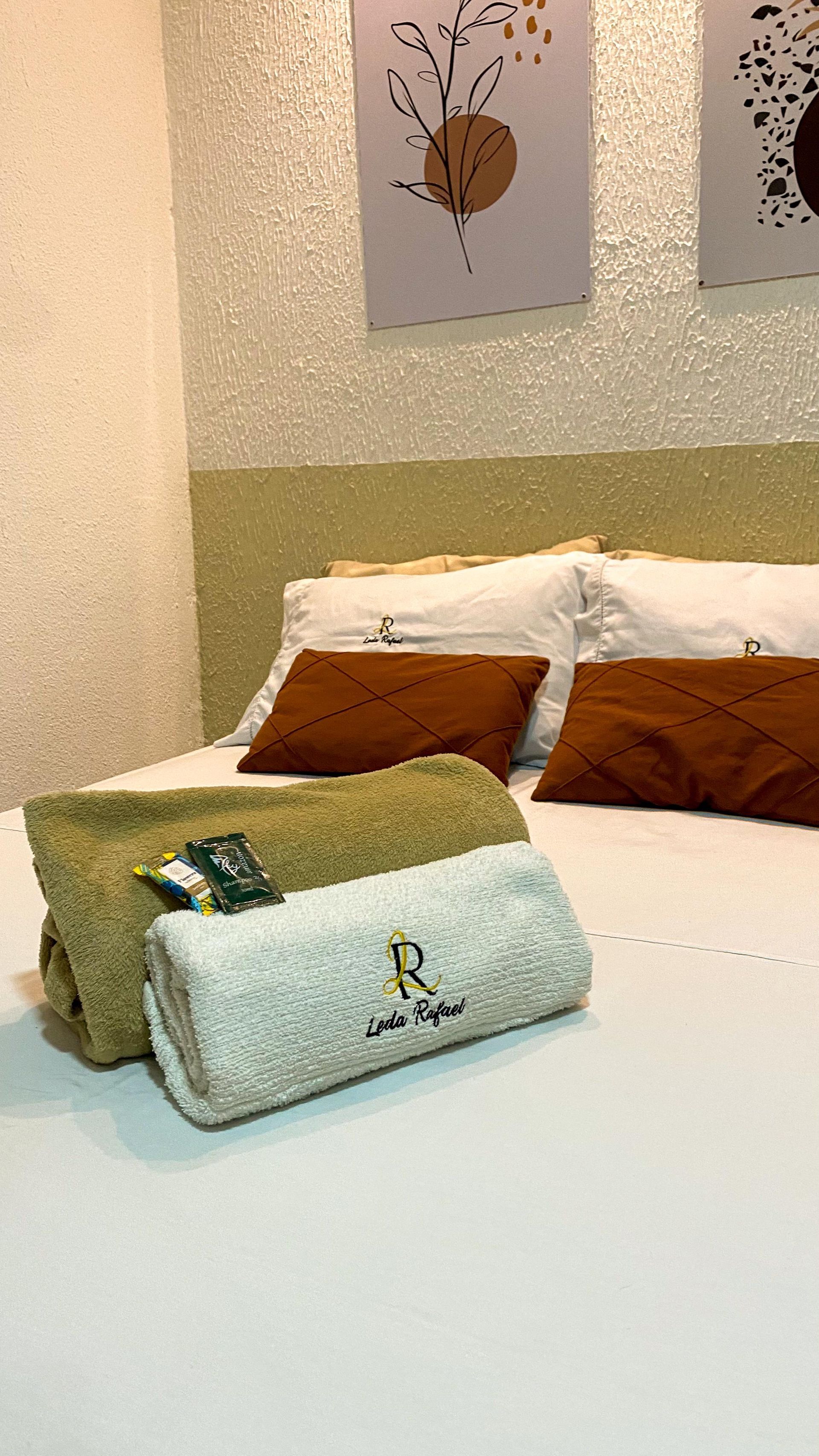Cama com lençóis brancos, travesseiros marrons, toalhas enroladas verdes e brancas e cabeceira bege encostada em uma parede texturizada com obras de arte.