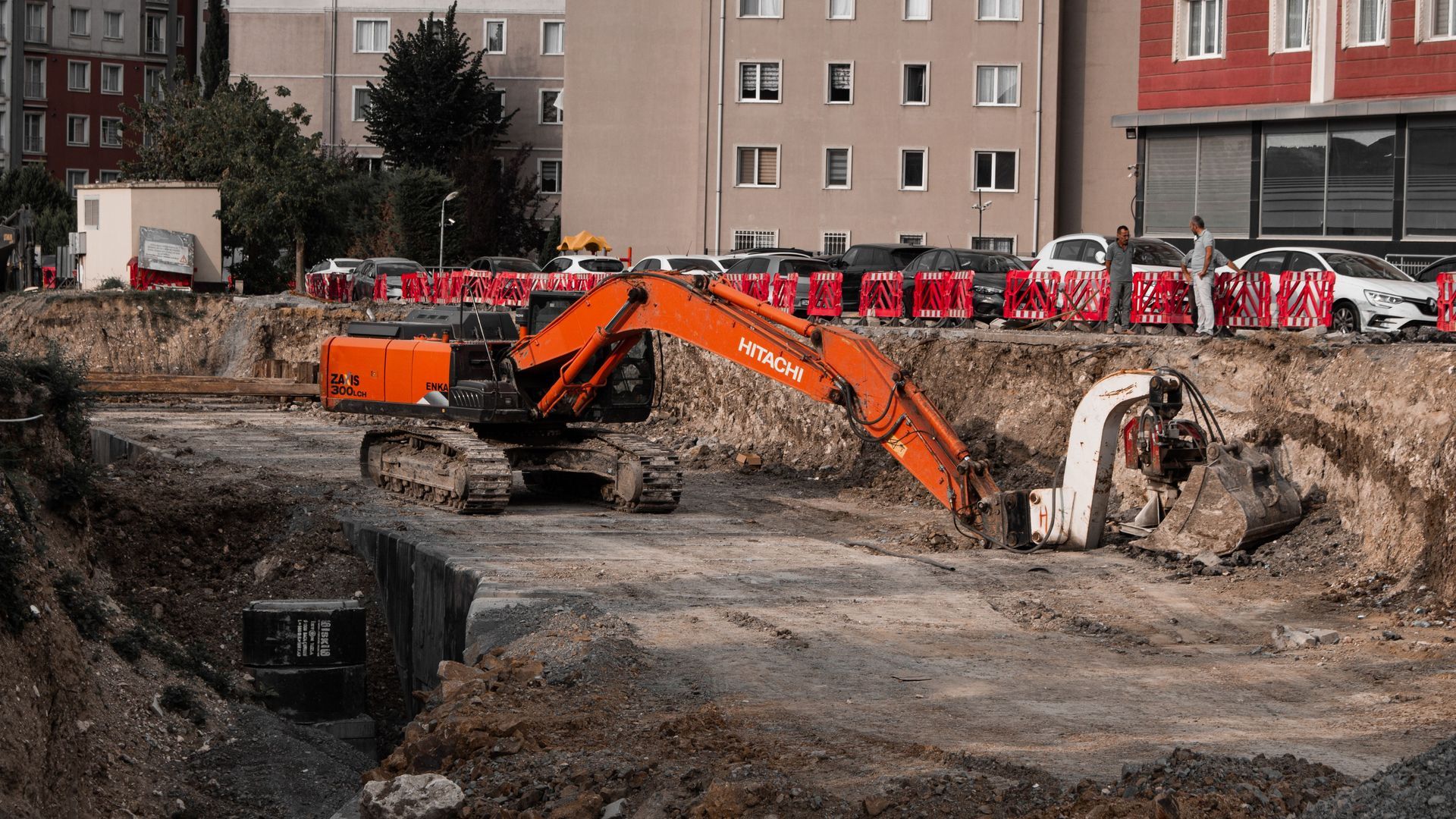 Orange excavator digging a large construction pit beside apartment buildings and parked cars