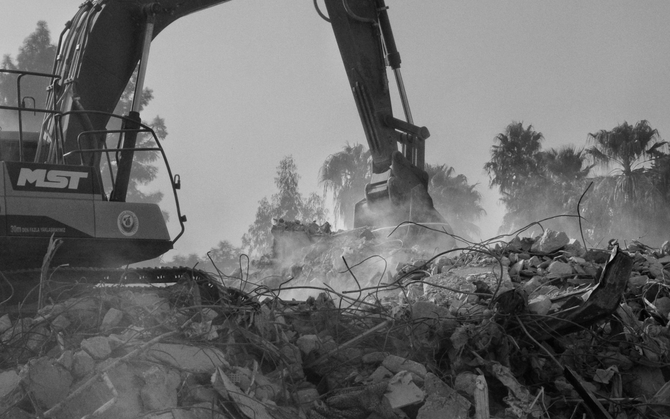 Excavator demolishing rubble amid dust and debris, with another machine and trees in the background