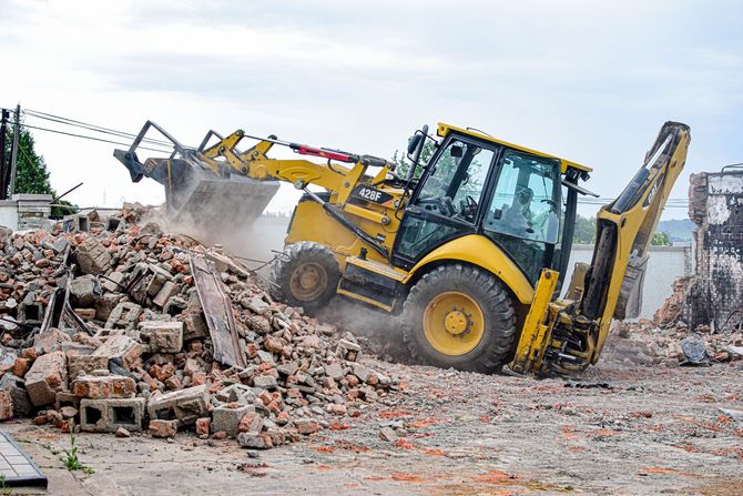 Yellow backhoe loader demolishing a pile of rubble at a construction site with dust flying