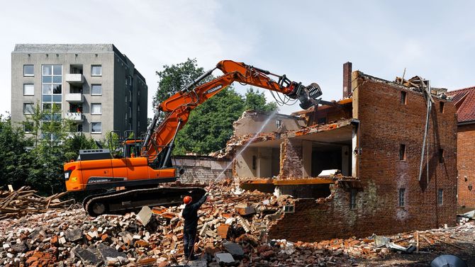 Orange excavator demolishing a brick building amid rubble, with a gray apartment block in the background