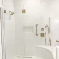White tiled shower with built-in shelf, grab bar, showerhead, and handheld sprayer.