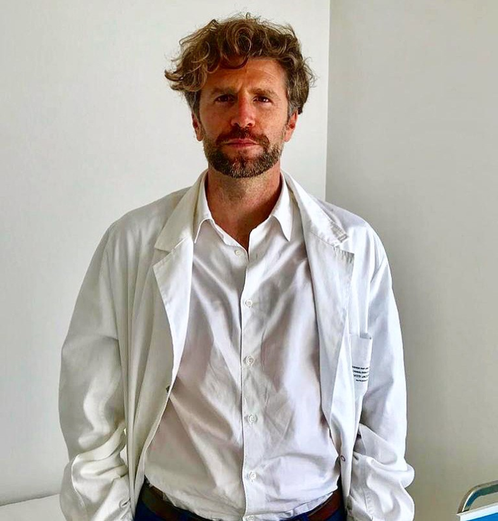 Uomo in piedi davanti a una parete spoglia, con una camicia bianca e una giacca bianca, dall’espressione seria.