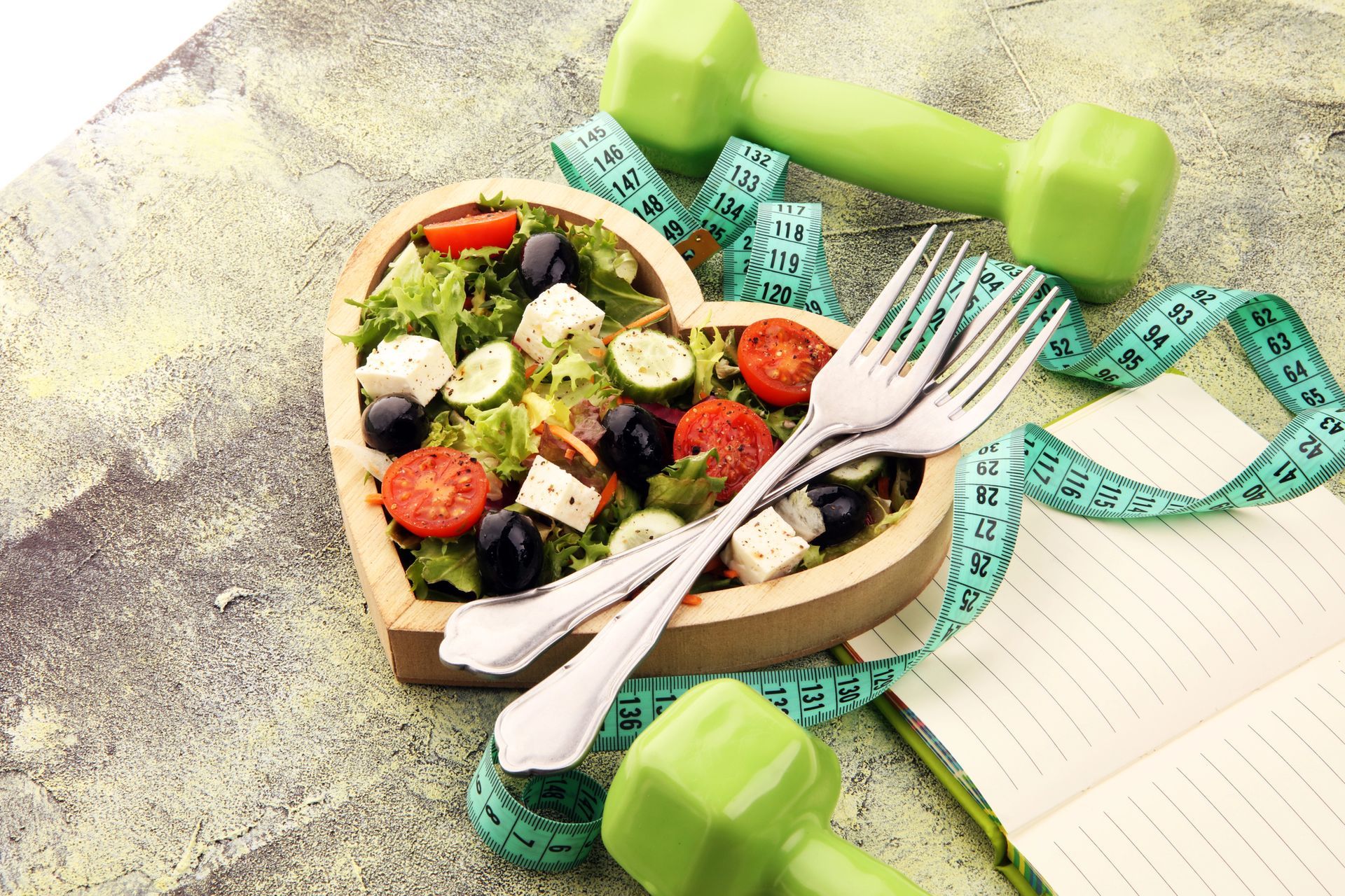 Pranzo sano in un contenitore con insalata, forchette, quaderno, metro a nastro e manubri su una superficie chiara