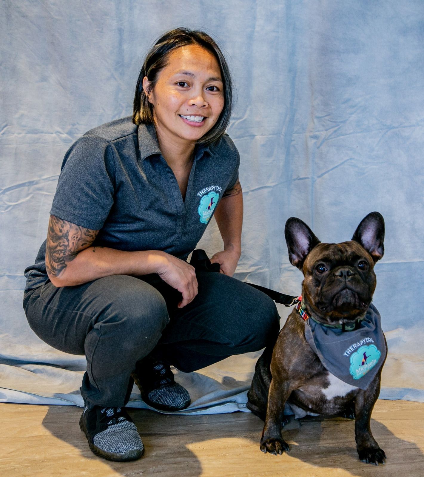 A smiling professional in a grey uniform crouches next to a brown French bulldog wearing a matching grey bandana.