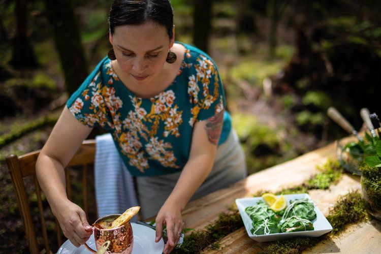 A woman is sitting at a table in the woods preparing food.