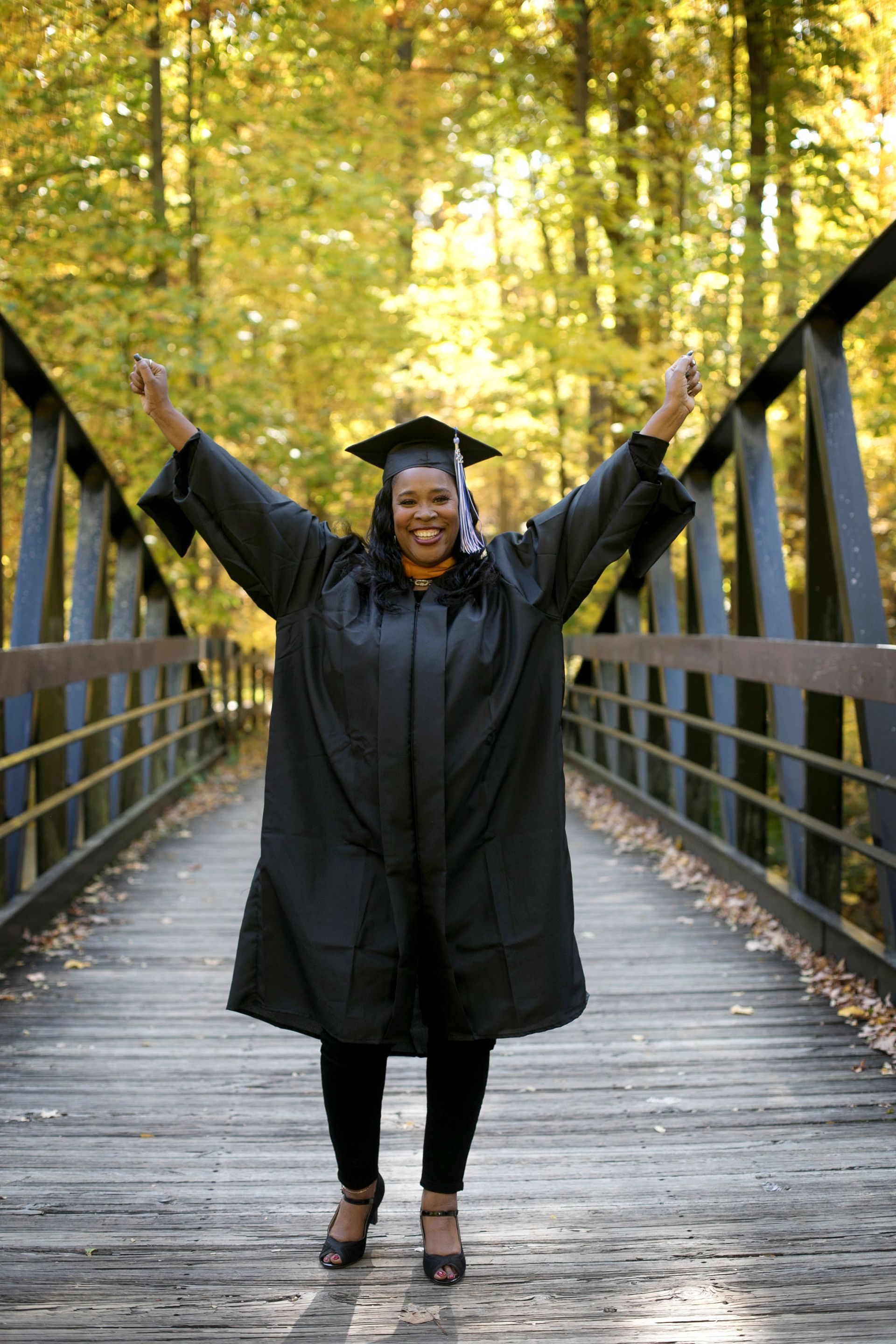 Photo of Maria Rush in graduation gown and cap on bridge with arms raised in celebration