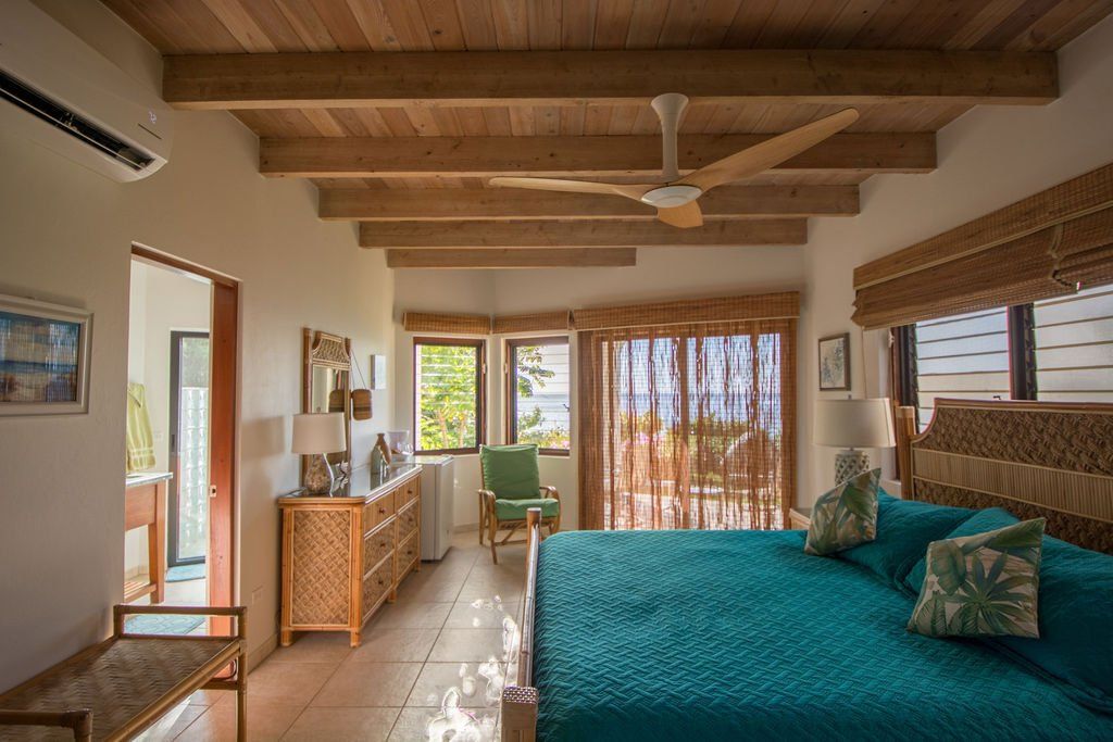 Bright bedroom with teal bed, wooden ceiling beams, ceiling fan, and large windows with sheer curtains