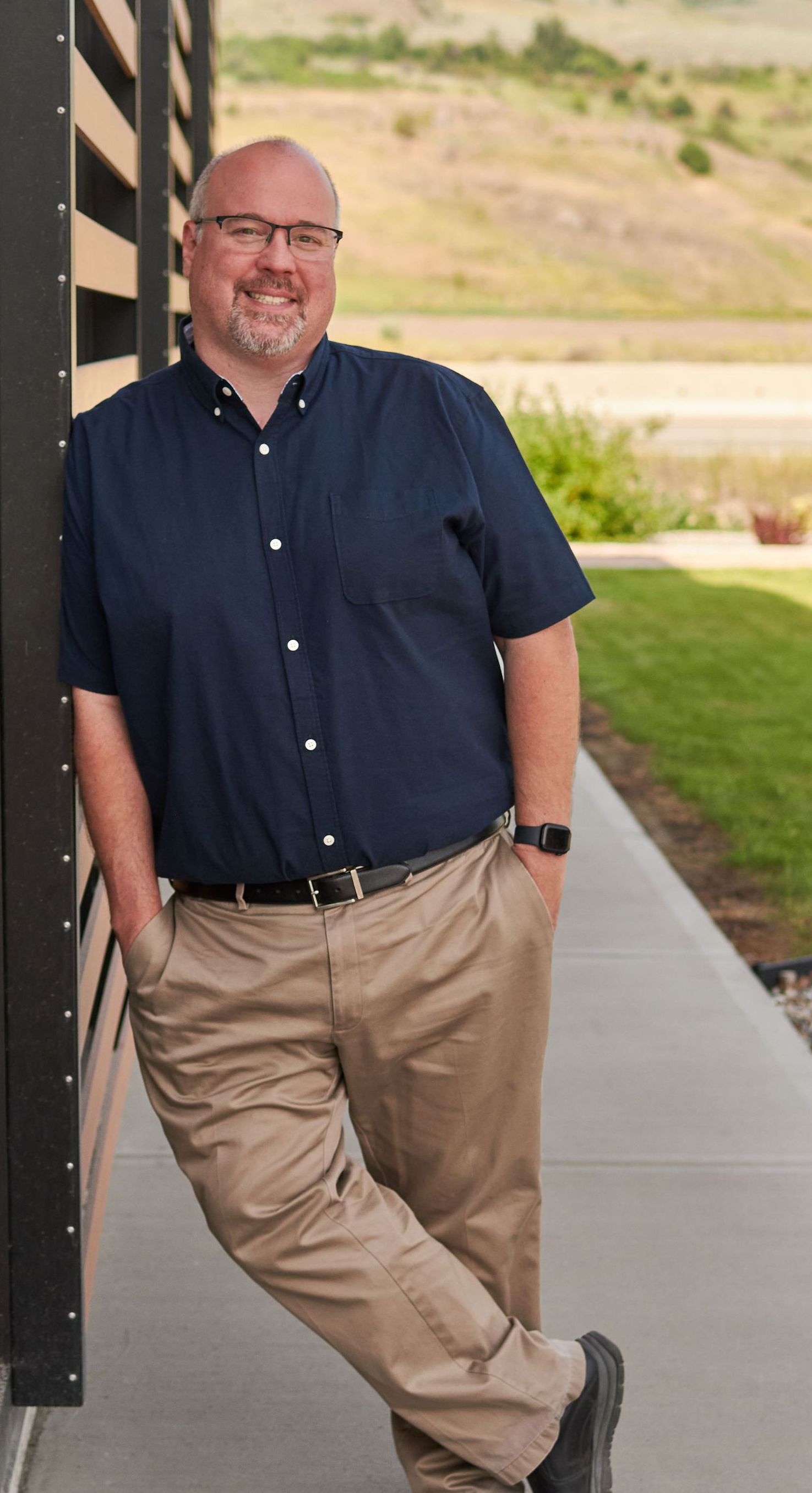 A handsome middle aged man in a blue shirt and khaki pants is leaning against a wall giving the impression of being trustworthy and reliable.