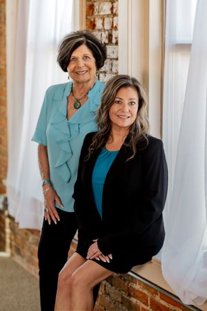 Two women posing by a brick wall and window, one seated in a black blazer and one standing in a teal blouse.
