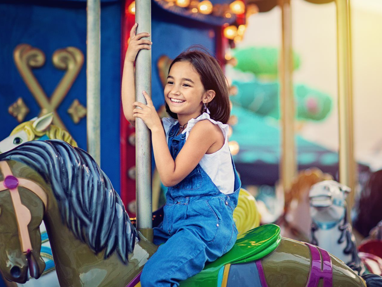 Child smiling on a colorful carousel horse, holding a pole at an amusement park.