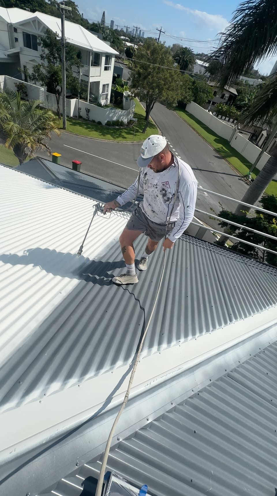 Man painting a corrugated metal roof white, using a roller. Safety rope attached.