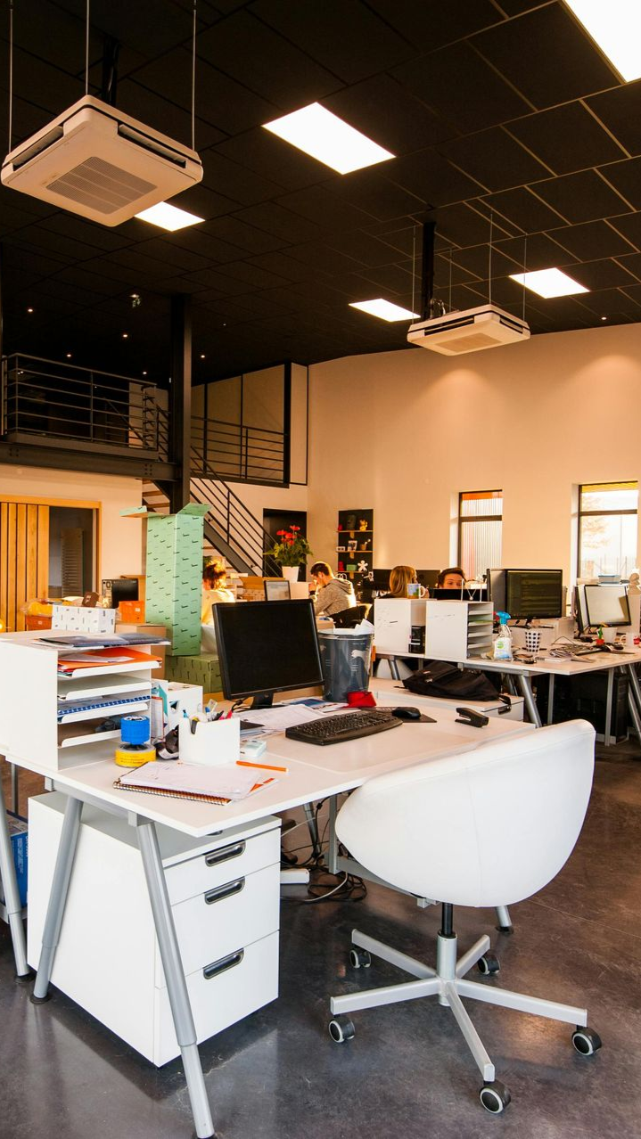 Office interior with desks, chairs, and multiple computers. Black ceiling, white walls, and a staircase.