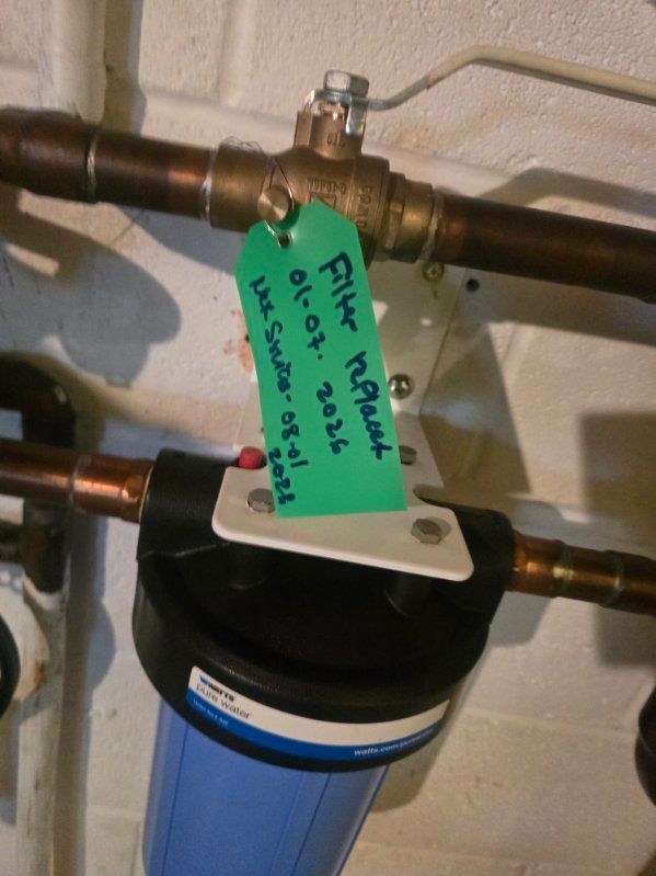 Blue water filter tank under copper pipes with a brass valve and green handwritten tag.
