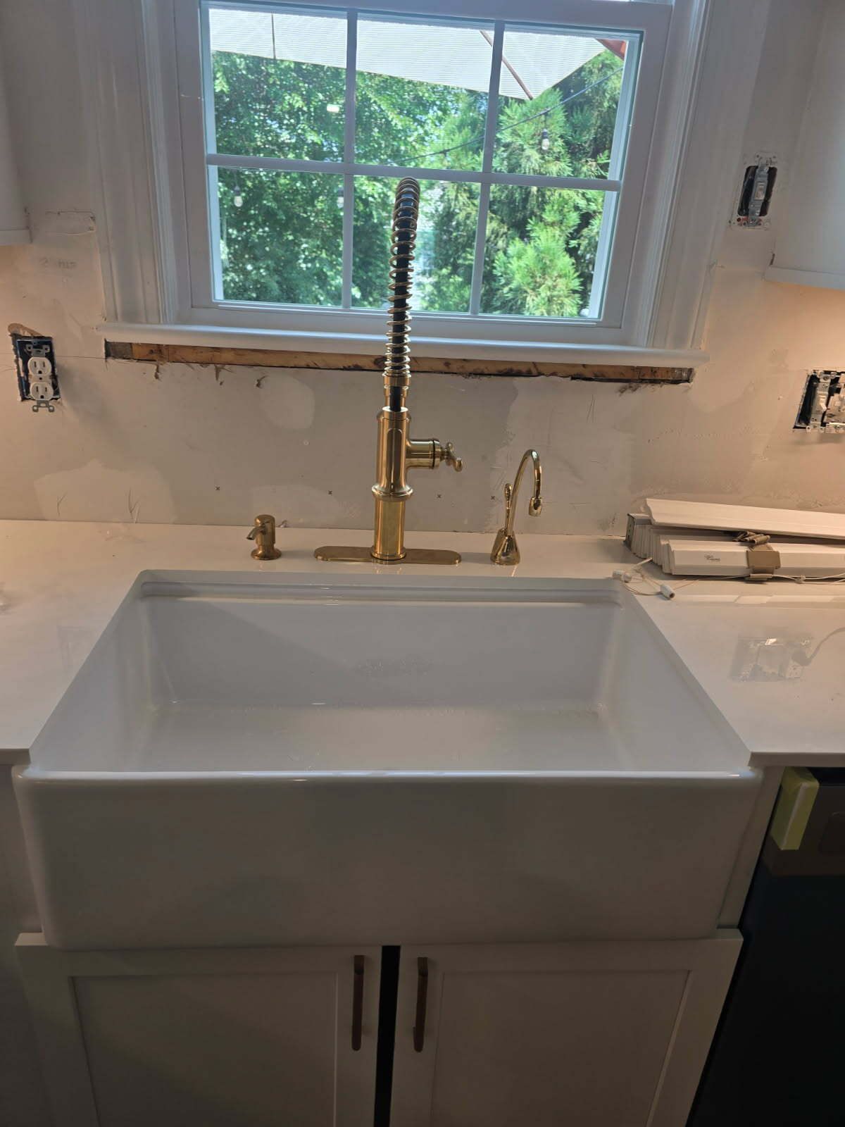 White bathroom vanity with a square sink and gold faucet beneath a window with greenery outside
