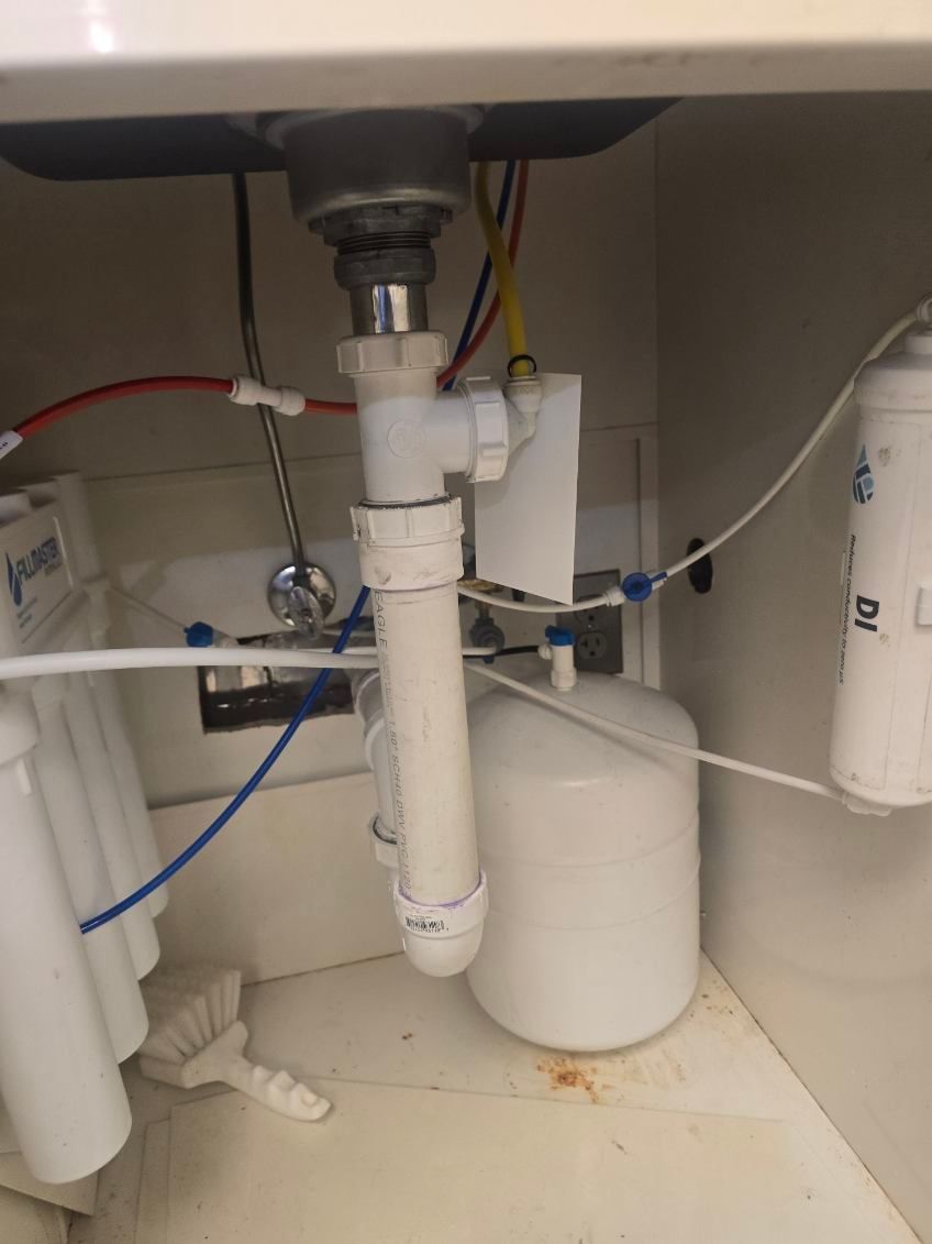 Under-sink plumbing with white PVC pipes, hoses, and two filter canisters inside a cabinet