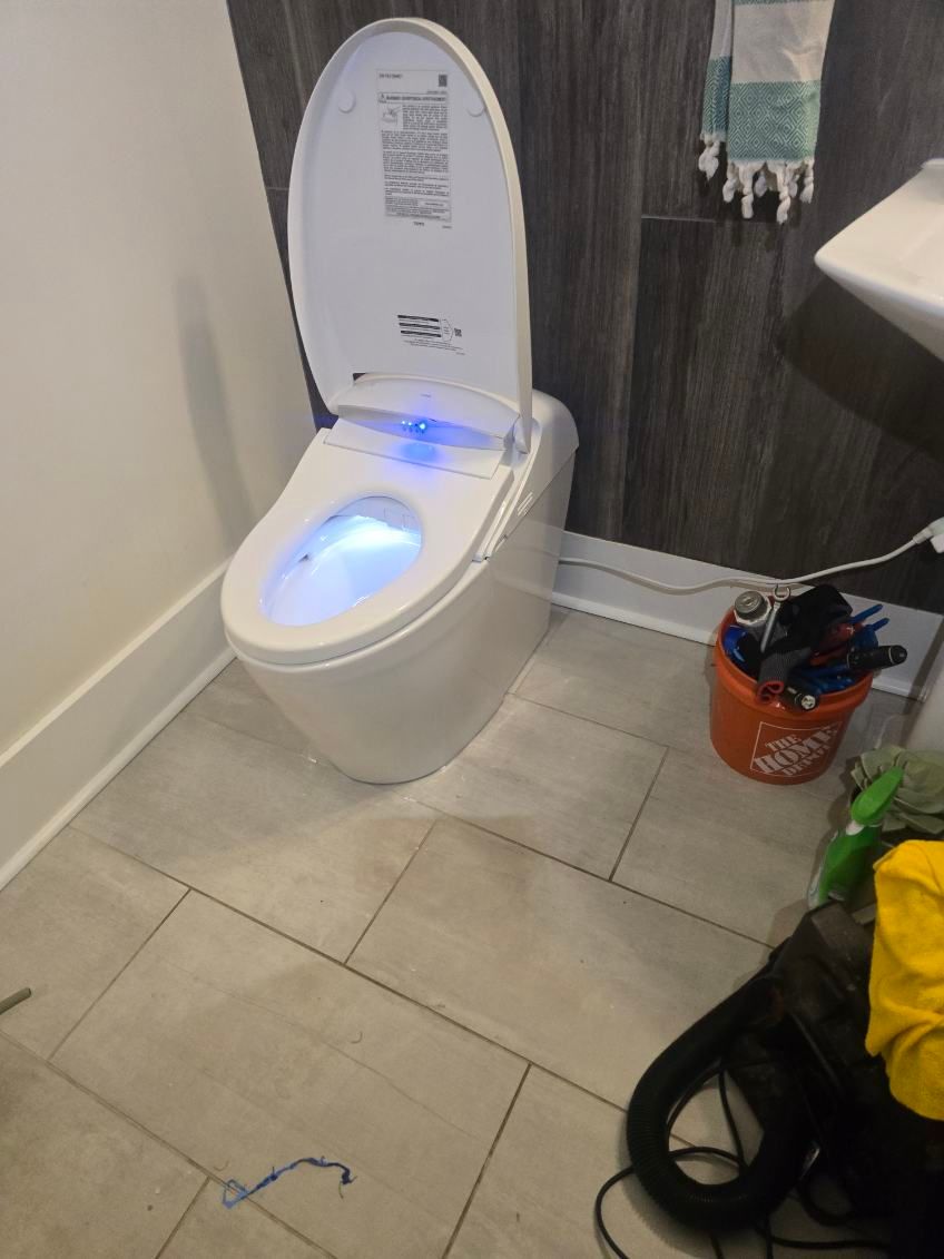 Bathroom with a lit toilet, open lid, tile floor, and cleaning supplies nearby