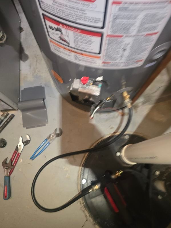 Water heater with tools and hoses on a basement floor, suggesting maintenance work.