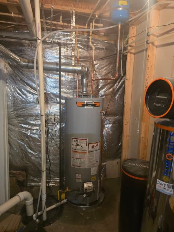 Basement utility room with a water heater, pipes, insulation, and a vacuum hose on the right.