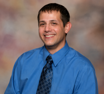 Man in blue shirt and patterned tie smiles; blurred background.