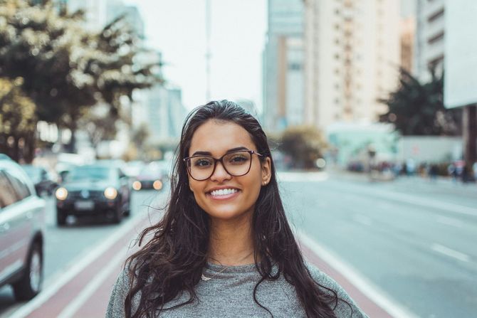 Woman with glasses smiles on a city street, cars and buildings in background.