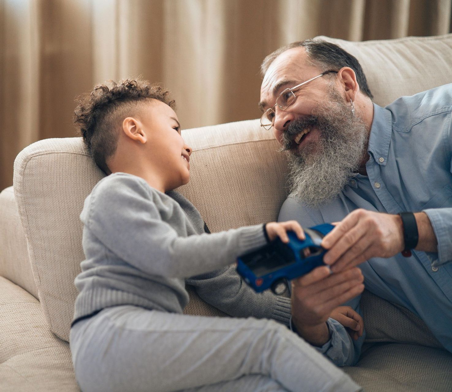 Grandfather giving a toy car to a smiling young boy, both sitting on a couch.