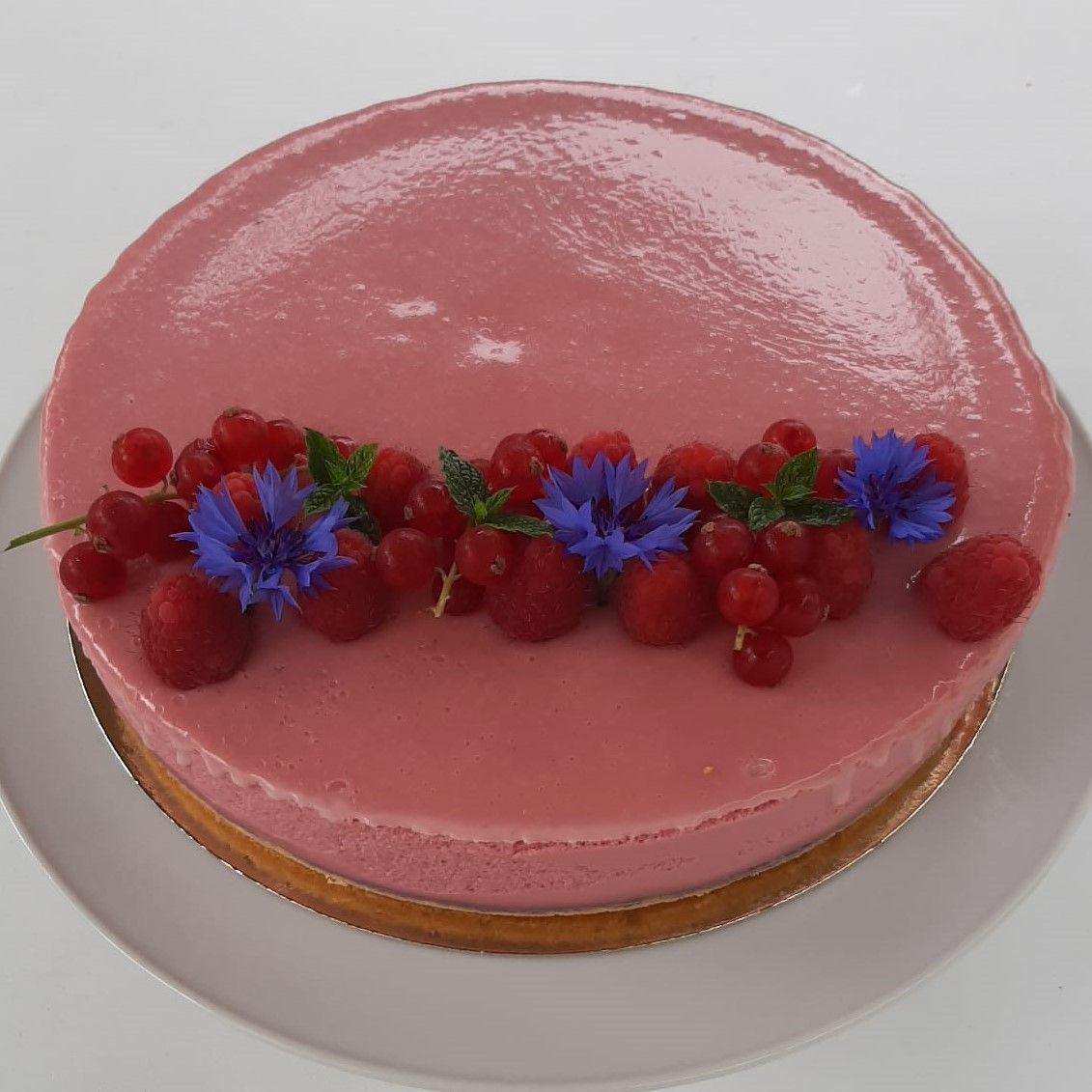 Roze geglazuurde taart, versierd met rode bessen, munt en blauwe bloemen, op een wit bord.