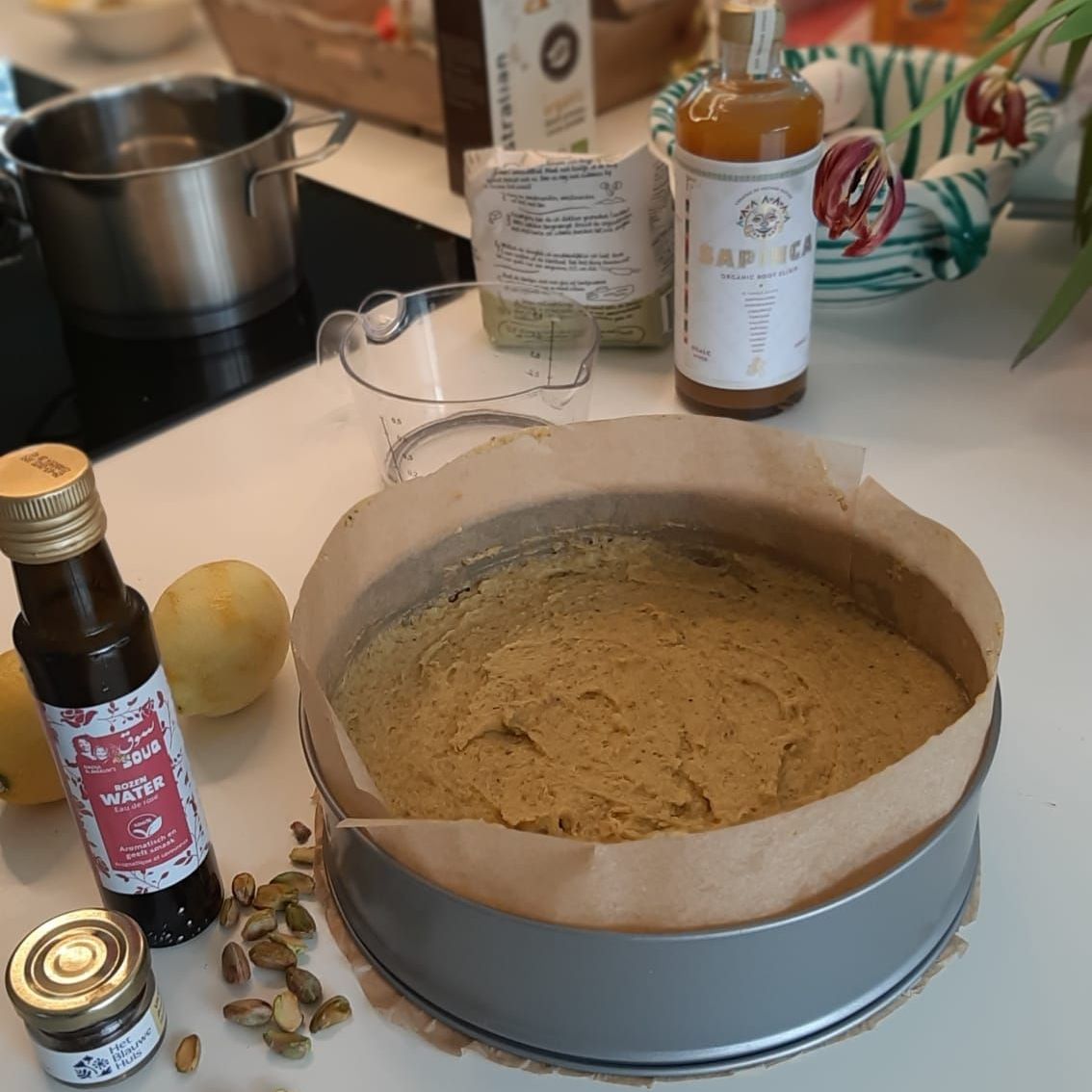 Cakebeslag in een bakvorm, klaar om te bakken, met ingrediënten zoals olie, siroop, noten en citroenen op het aanrecht.