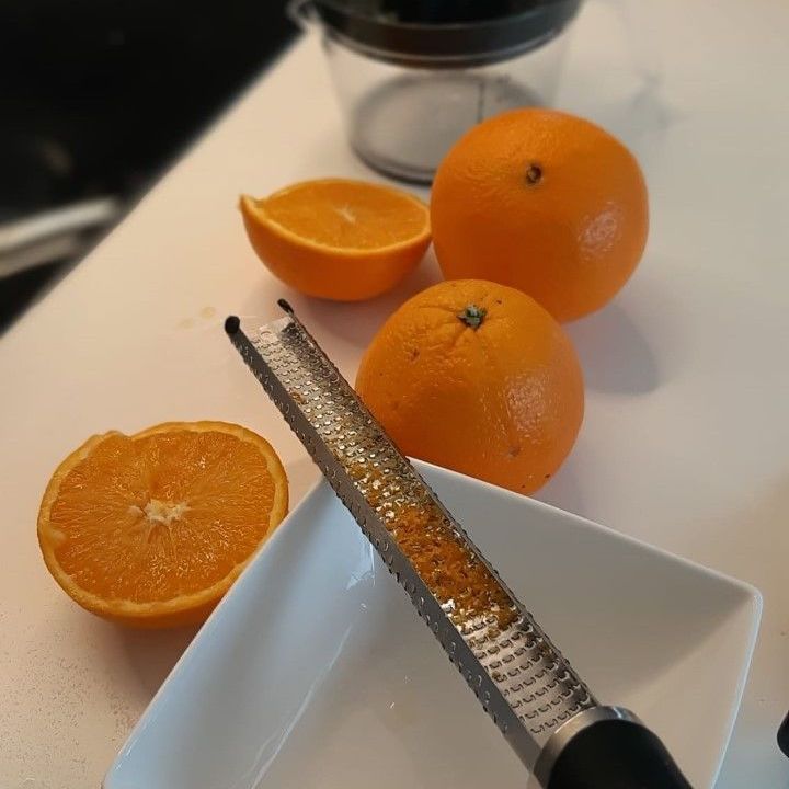 Sinaasappelen, een halve sinaasappel, een rasp, de rasp van de sinaasappelschil en een witte kom op een witte ondergrond