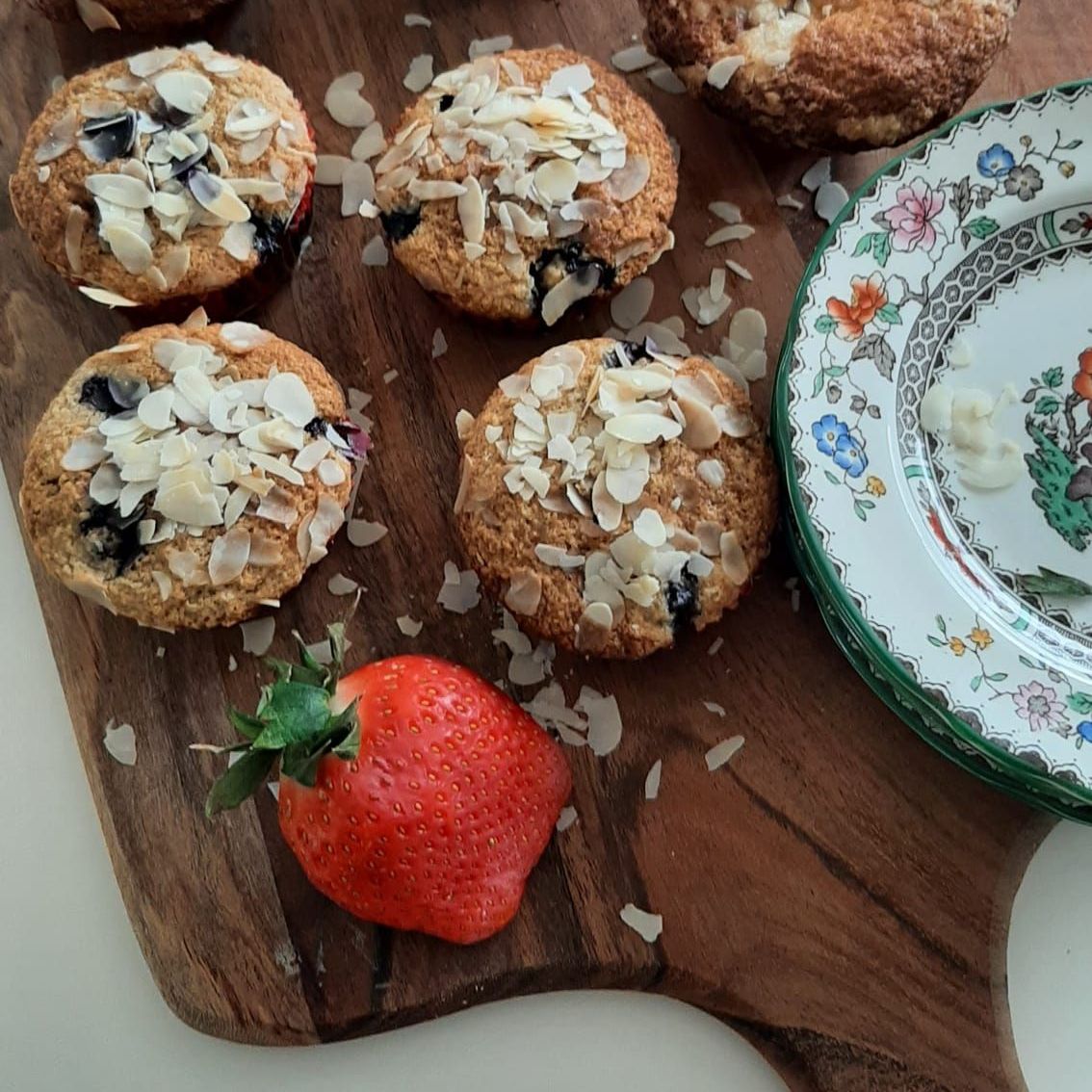 Bosbessenmuffins met amandelschilfers, aardbeien en een bloemmotief op een houten plank.