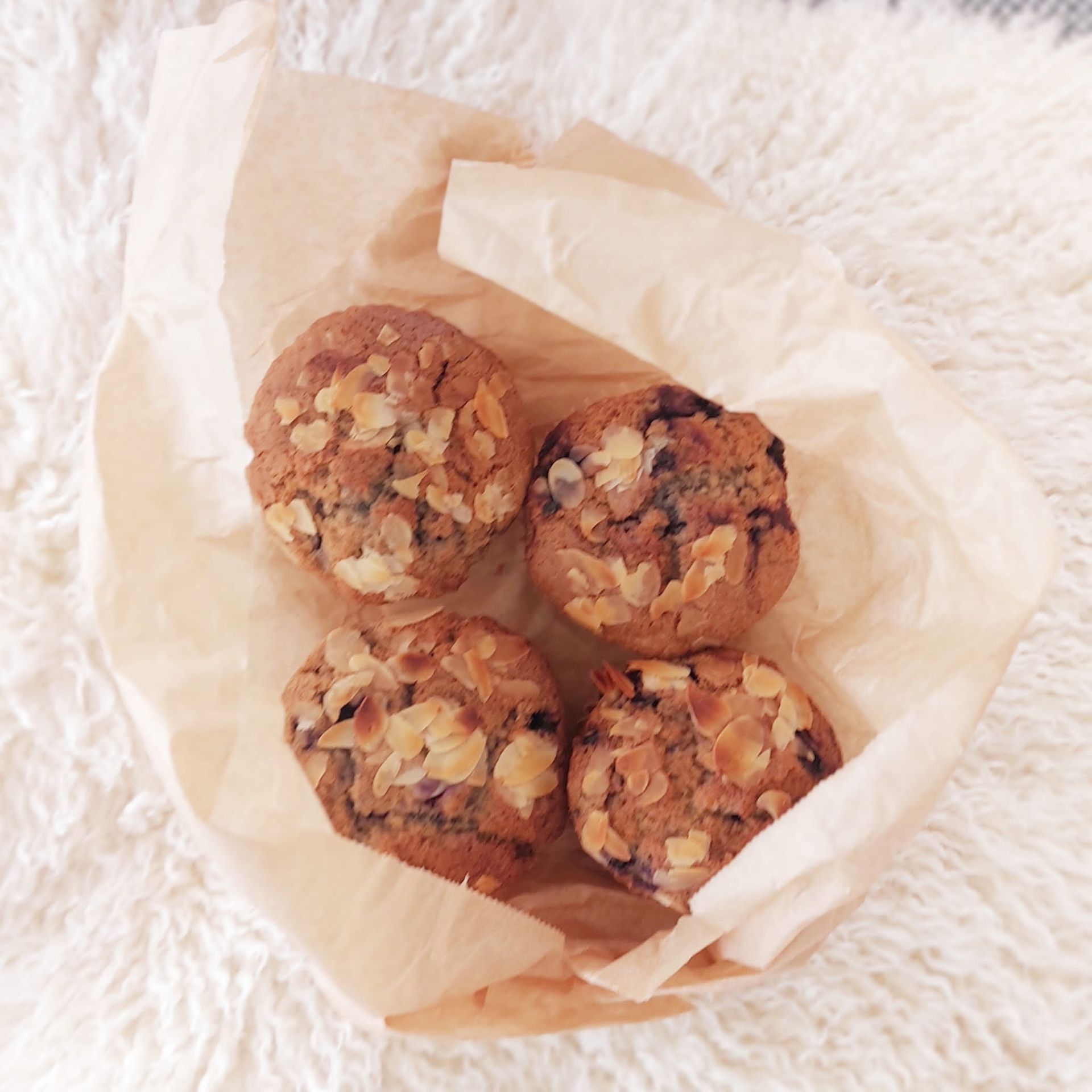 Vier muffins in een bruine papieren zak, bedekt met amandelen, op een witte, luchtige ondergrond.