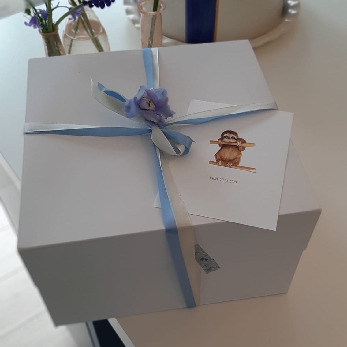 Witte geschenkdoos, vastgebonden met een lichtblauw lint, en een kaartje met een illustratie van een luiaard.