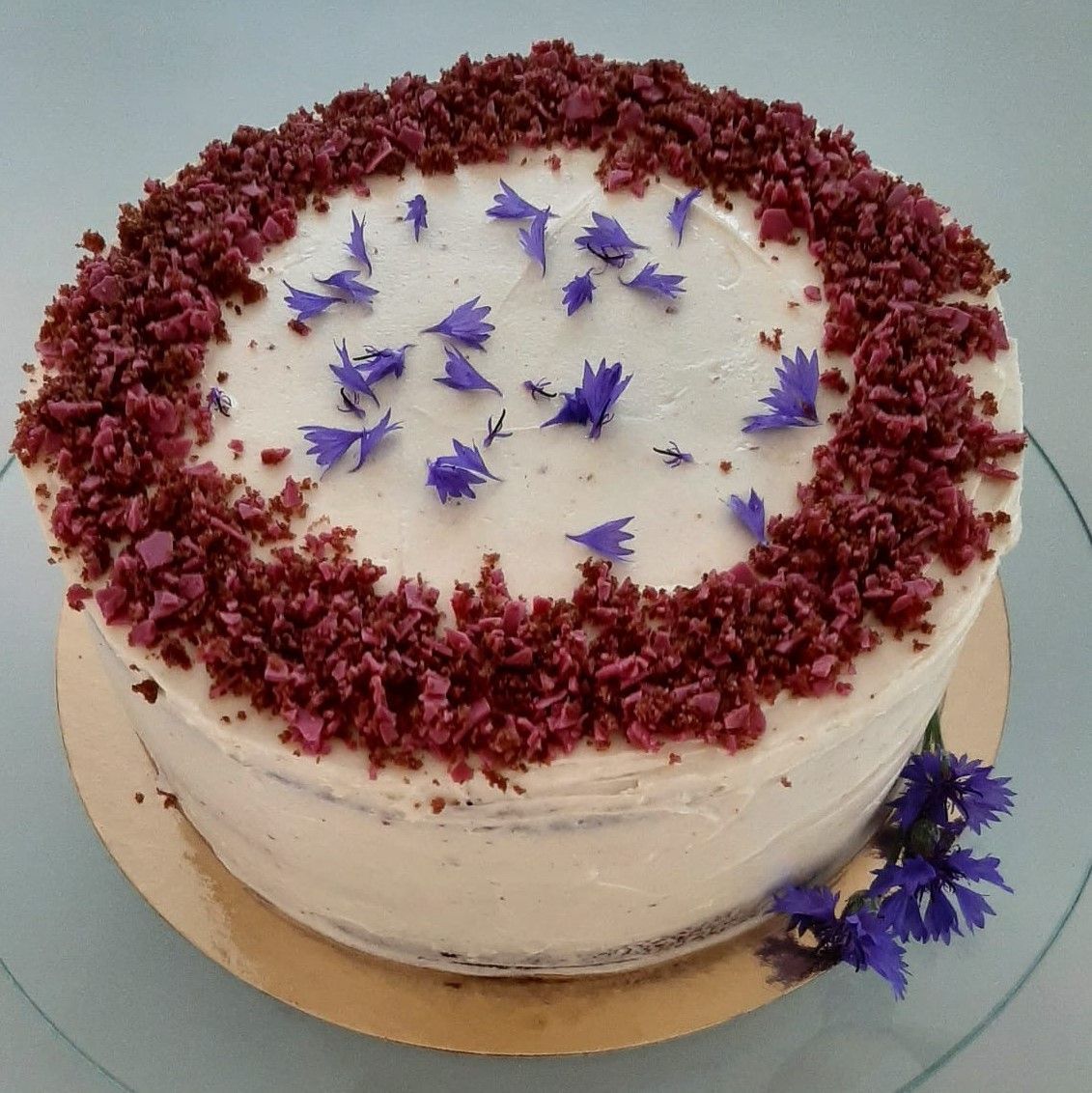 Taart met witte glazuur, afgewerkt met bordeauxrode kruimels en versierd met blauwe bloemen.
