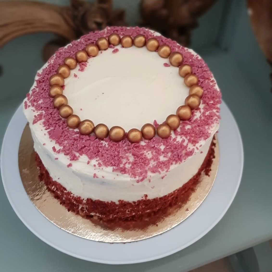 Rode fluwelen cake met witte glazuur, roze hagelslag en een gouden parelrand op een wit bord.