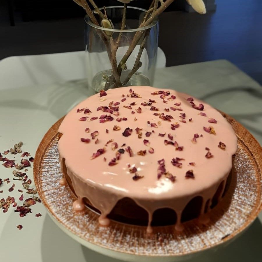 Taart met roze glazuur en rozenblaadjes, op een decoratief bord, met een vaas met bloemen op de achtergrond.