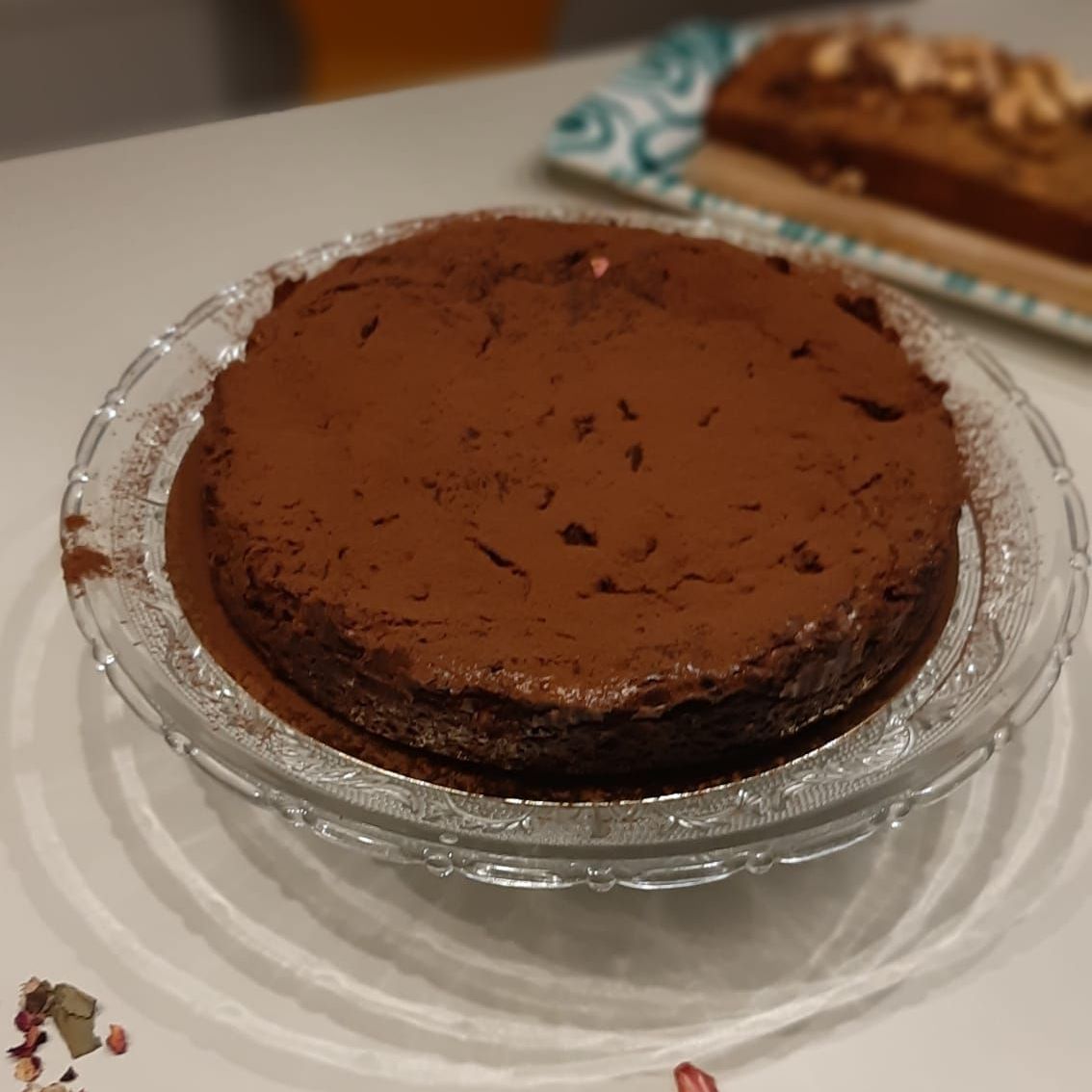 Chocoladetaart bestrooid met cacaopoeder op een glazen bord, met op de achtergrond nog een taart.