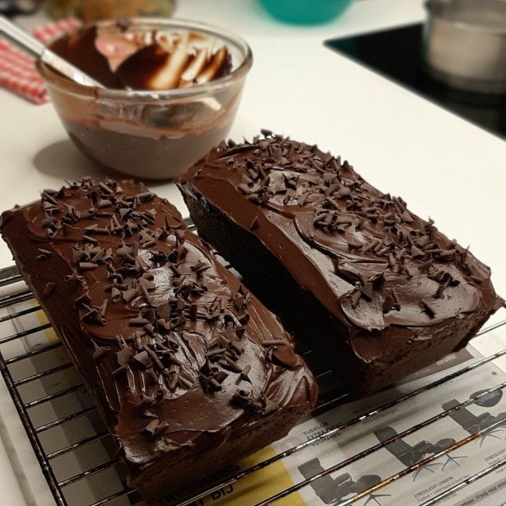 Twee chocoladebroden, bedekt met chocoladeglazuur en chocoladeschaafsel, staan op een rooster, met het glazuur in een kom.