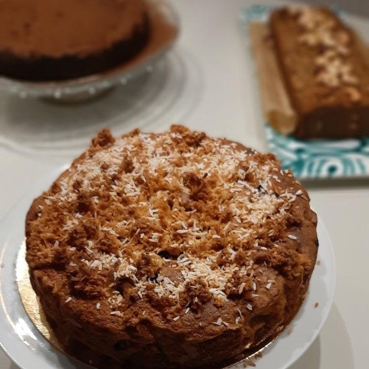 Een ronde taart met een gestructureerde topping en geraspte kokos. Op de wazige achtergrond zijn andere taarten te zien.