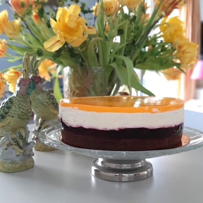 Taart met lagen bruin, wit en geel, bedekt met gelei, op een glazen taartplateau, met tulpen op de achtergrond.