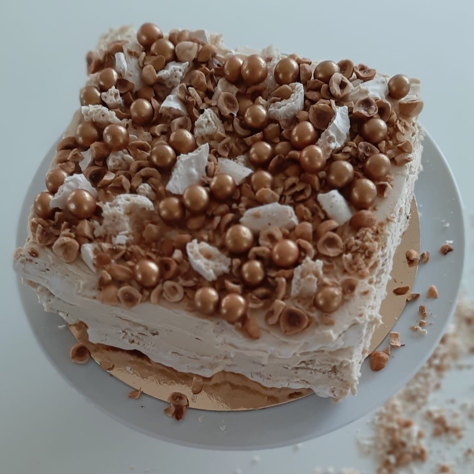 Vierkante taart met een lichte glazuurlaag, versierd met goudkleurige bolletjes, gehakte noten en stukjes meringue.